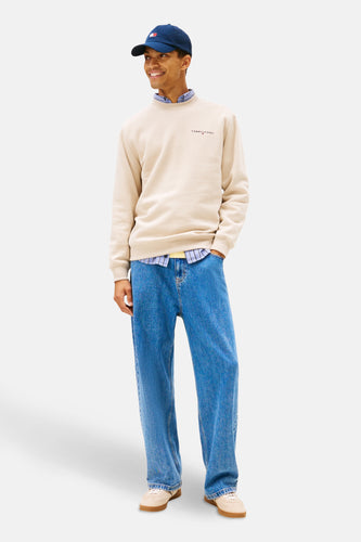 Een man staat, gekleed in een beige trui, blauw overhemd, blauwe jeans en een blauwe pet.