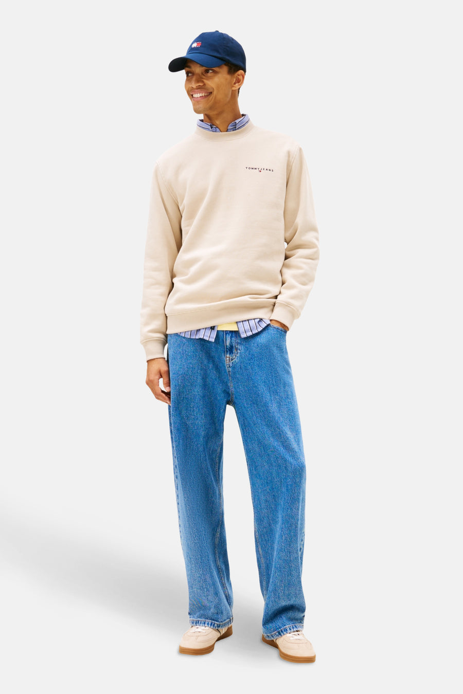 Een man staat, gekleed in een beige trui, blauw overhemd, blauwe jeans en een blauwe pet.