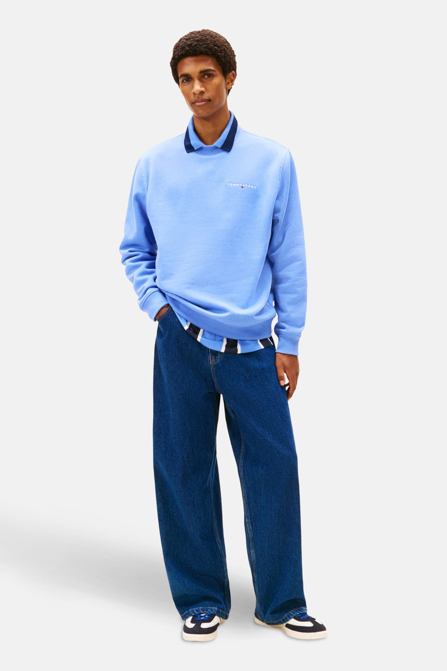 Een man die een lichtblauwe trui, blauwe jeans en zwarte schoenen draagt.