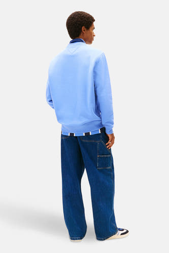 Pull-over bleu de TOMMY JEANS, avec des manches longues, associé à des jeans larges et des baskets basses.