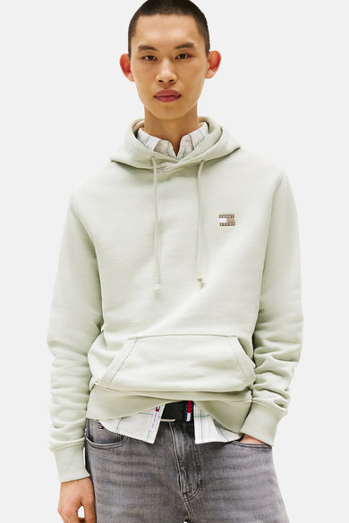Pull-over vert de TOMMY JEANS avec capuche, porté sur une chemise à carreaux et un ceinture noire.