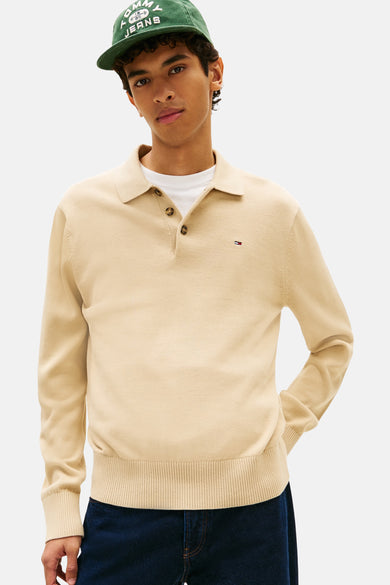 Pull beige de TOMMY JEANS, à manches longues, col polo, avec un t-shirt blanc en dessous et un jean bleu.