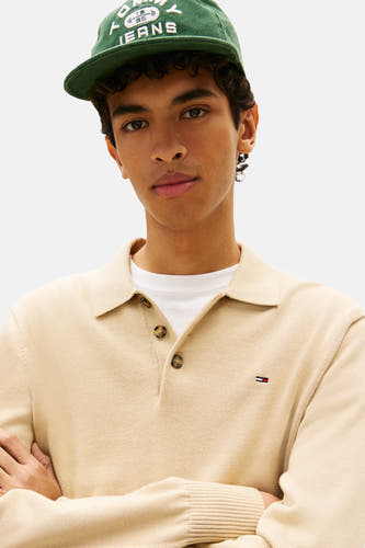 Pull beige de TOMMY JEANS, avec col et logo sur la poitrine, porté sur une chemise blanche et avec une casquette verte.