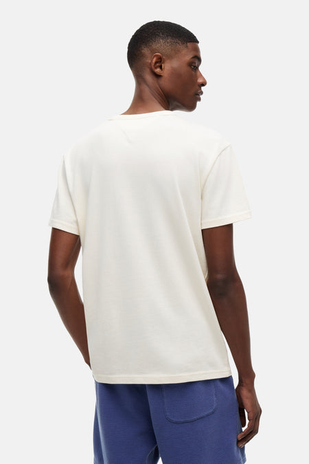 T-shirt à manches courtes de couleur crème de TOMMY JEANS, dos visible avec coutures en V au niveau du cou, porté avec un short gaufré bleu.