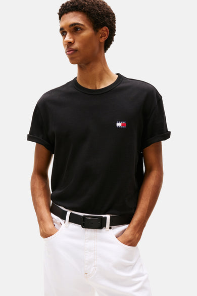 T-shirt noir à manches courtes de TOMMY JEANS, avec des manches retroussées et un petit logo sur la poitrine, porté avec un pantalon blanc et une ceinture noire.
