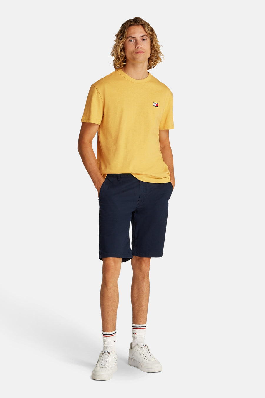 T-shirt jaune à manches courtes de TOMMY JEANS, porté avec des shorts bleus, des chaussettes blanches et des baskets.
