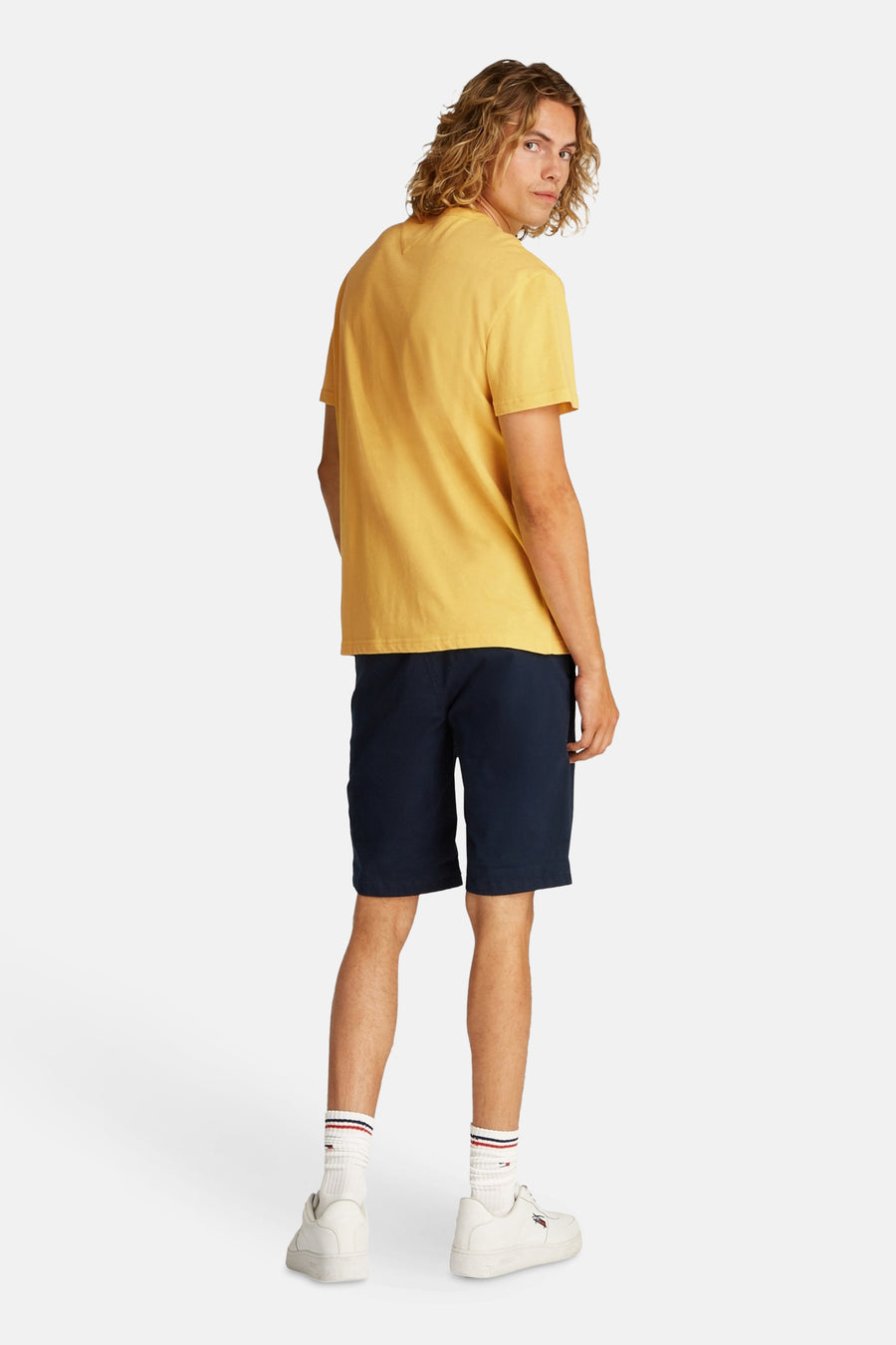 Geel T-shirt met korte mouwen van TOMMY JEANS, gezien van achteren, met donkerblauwe shorts, witte sokken en sneakers.