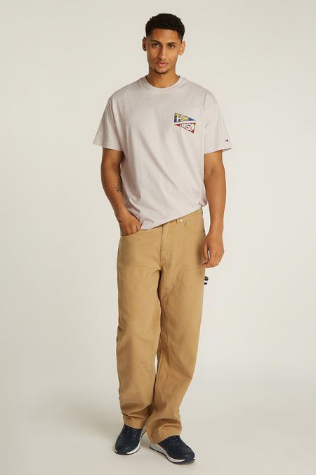 T-shirt écru à manches courtes de TOMMY JEANS, avec un logo sur la poitrine et porté avec un pantalon marron et des baskets bleues.