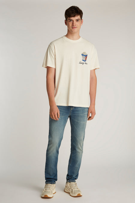 T-shirt blanc à manches courtes de Tommy Jeans, avec imprimé popcorn sur la poitrine, associé à des jeans bleus et des baskets crème.