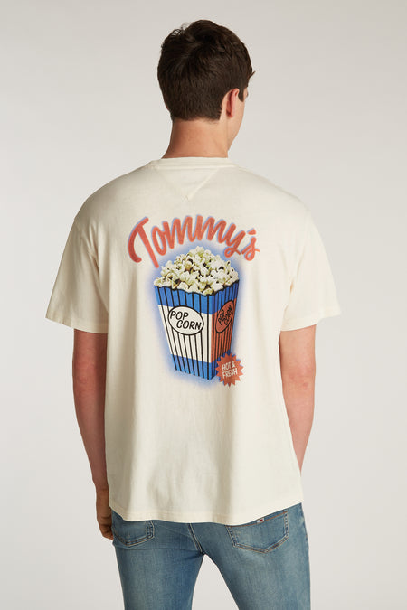 T-shirt blanc à manches courtes de TOMMY JEANS, avec une image de popcorn sur le dos et porté avec un jean.