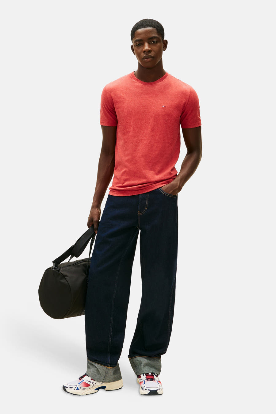 Rood T-shirt met korte mouwen van TOMMY JEANS, gedragen met donkerblauwe wijde jeans en sportschoenen, met een zwarte tas.
