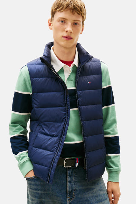 Gilet bleu de TOMMY JEANS porté sur un t-shirt rayé et des jeans larges avec des chaussures marrons.