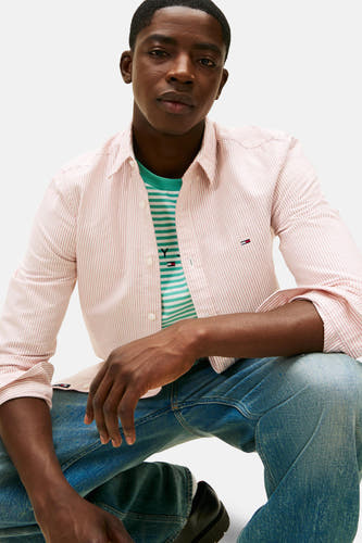 T-shirt rayé rouge de TOMMY JEANS avec des manches longues, doté d'un logo brodé, porté sur un t-shirt rayé et des jeans.