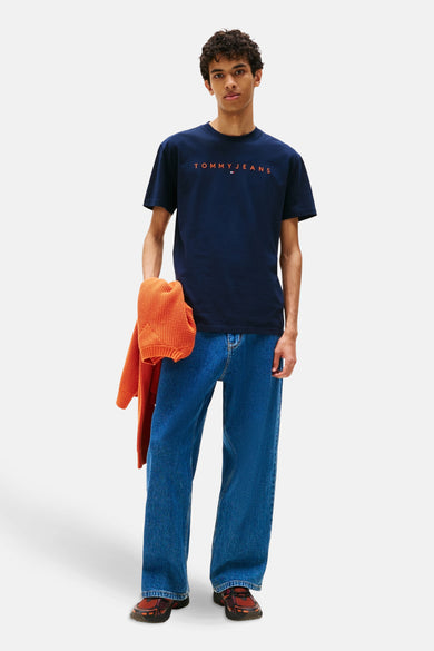 T-shirt bleu à manches courtes de TOMMY JEANS, avec un logo orange et porté avec un gilet orange et des jeans bleus.