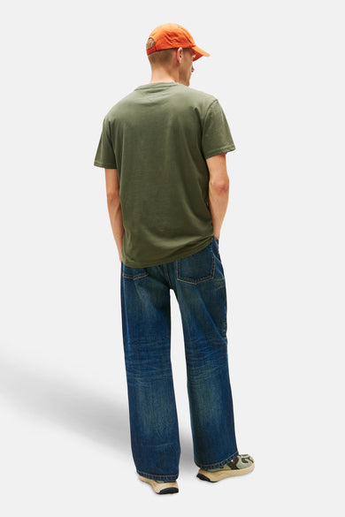 T-shirt vert avec manches courtes de TOMMY JEANS, associé à des jeans larges et une casquette orange.