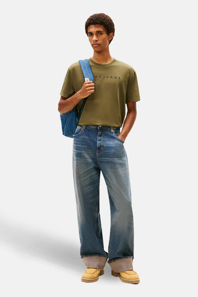 Groen T-shirt met korte mouwen van TOMMY JEANS, met een blauwe denim rugzak over de schouder en wijde jeans.
