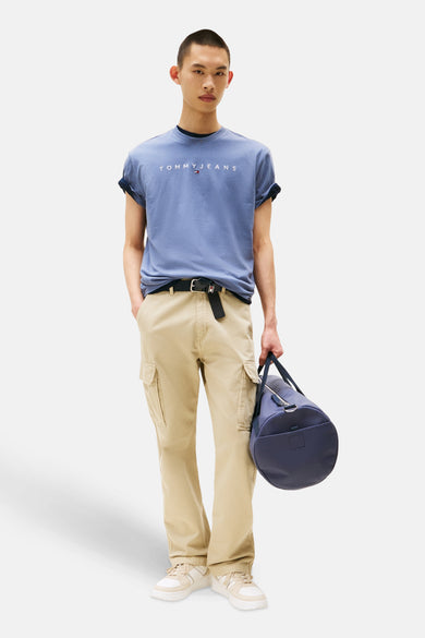 T-shirt bleu à manches courtes de TOMMY JEANS, avec des manches retroussées, porté avec un pantalon kaki, une ceinture et un sac marine.