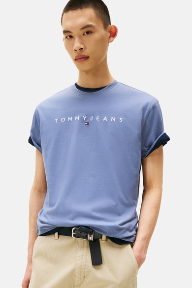 T-shirt bleu à manches courtes de TOMMY JEANS, avec des manches retroussées et un logo sur la poitrine, associé à un pantalon marron et une ceinture noire.