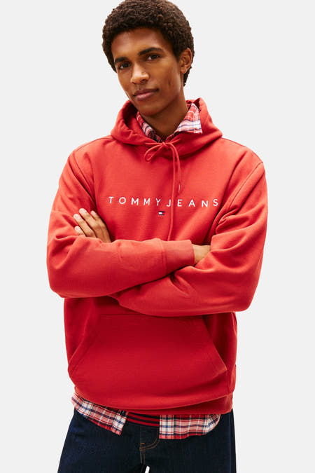 Pull-over rouge avec capuche de TOMMY JEANS, porté sur une chemise à carreaux et associé à des jeans bleu marine larges.