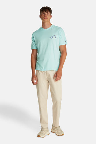 T-shirt turquoise à manches courtes de TOMMY JEANS, avec le nom de la marque brodé, porté avec un pantalon beige et des baskets.