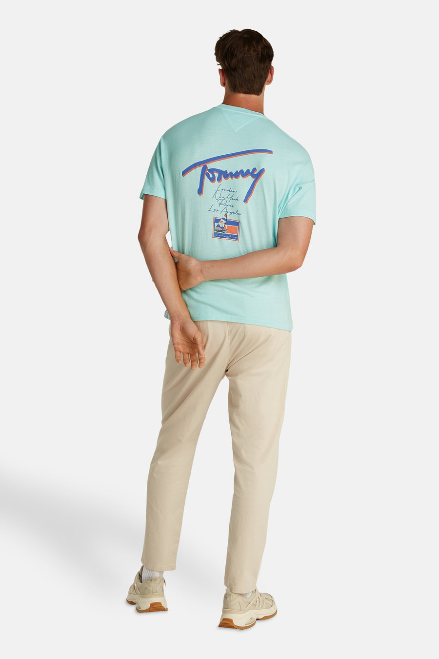 T-shirt turquoise à manches courtes de Tommy Jeans, dos imprimé avec le logo et des villes, porté avec un pantalon kaki et des baskets.