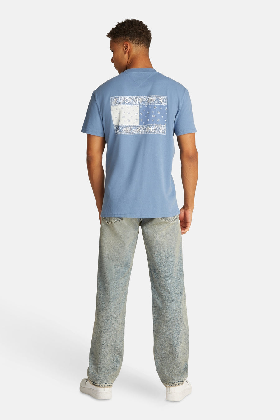 Man achteraan, blauw T-shirt, paisleyprint, wijde jeans, witte sneakers, lichtgrijze achtergrond.
