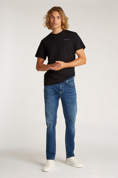 Zwart T-shirt met korte mouwen van TOMMY JEANS, gedragen met blauwe jeans en witte sneakers.
