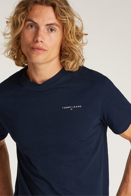 T-shirt bleu à manches courtes de TOMMY JEANS, porté par un homme aux cheveux blonds ondulés.