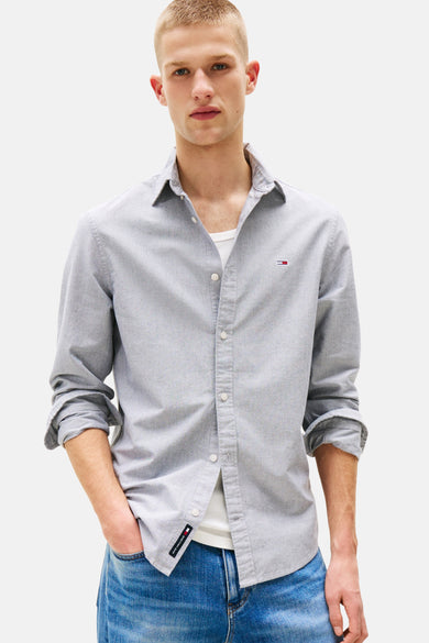 Chemise bleue à manches longues de TOMMY JEANS, avec col boutonné et manches retroussées portée sur un T-shirt blanc.
