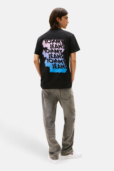 T-shirt noir à manches courtes de TOMMY JEANS, avec une impression de logo colorée sur le dos, associé à des jeans gris et des baskets blanches.