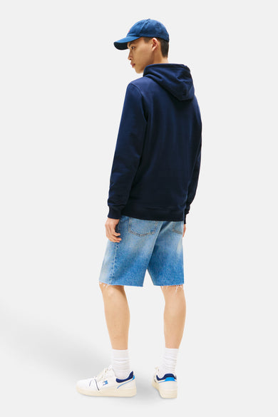 Blauwe sweater met kap van TOMMY JEANS, gezien van achteren, met denim shorts, witte sokken en sneakers.
