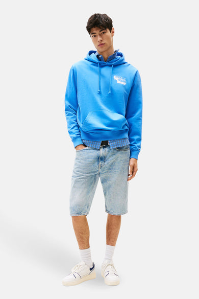 Pull-over bleu avec capuche de TOMMY JEANS, avec un logo blanc et associé à une chemise à carreaux et des shorts en denim.