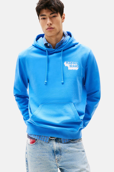 Pull-over bleu avec capuche de TOMMY JEANS, avec logo blanc, porté sur un chemisier à carreaux et des jeans.