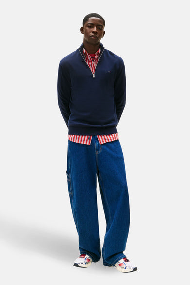Pull-over bleu de TOMMY JEANS, avec une fermeture éclair et porté sur un chemisier rayé, avec des jeans larges et des baskets.