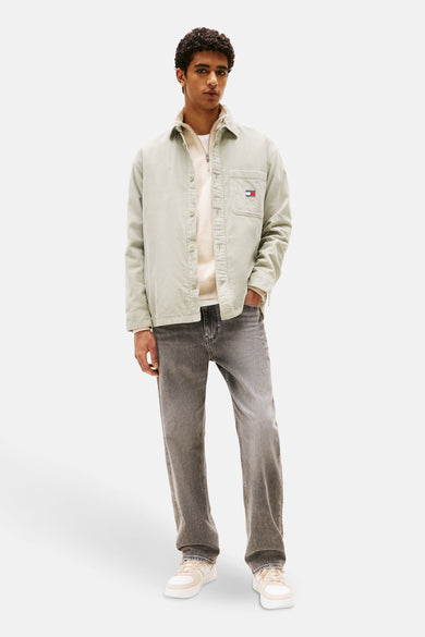 Chemise verte à manches longues de Tommy Jeans, avec une subtile texture corduroy. L'homme porte un pull blanc et des jeans gris.