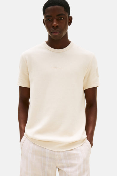 T-shirt beige à manches courtes de TOMMY JEANS, avec une subtile impression textuelle sur la poitrine et porté avec un short rayé.