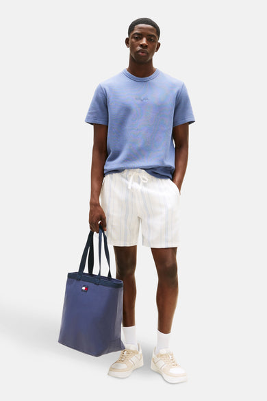 T-shirt bleu à manches courtes de TOMMY JEANS, porté avec des shorts rayés, des chaussettes blanches, des baskets basses et une sacoche bleue.