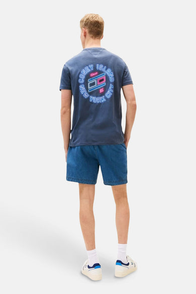 T-shirt bleu à manches courtes de TOMMY JEANS, avec une impression ronde sur le dos et porté avec des shorts en denim.