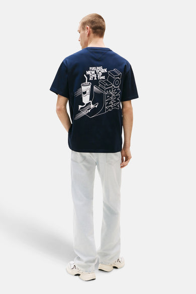 T-shirt bleu à manches courtes de TOMMY JEANS, avec une impression graphique sur l'arrière, porté avec des jeans bleus clairs et des baskets crème.