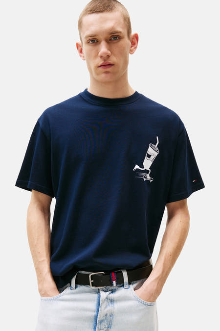T-shirt bleu à manches courtes de TOMMY JEANS, avec une impression blanche d'une tasse sur un skateboard, porté avec une ceinture et des jeans légers.