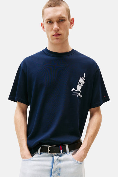 T-shirt bleu à manches courtes de TOMMY JEANS, avec une impression blanche d'une tasse sur un skateboard, porté avec une ceinture et des jeans légers.