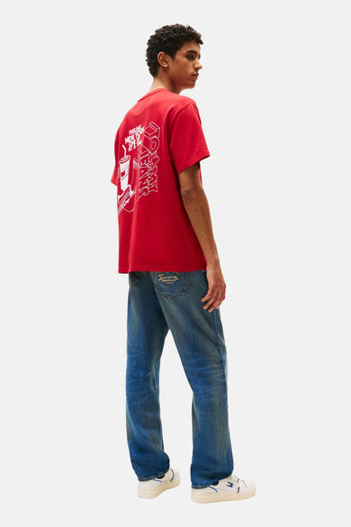 T-shirt rouge à manches courtes de Tommy Jeans, avec une impression blanche sur le dos, associé à des jeans bleus et des baskets blanches.
