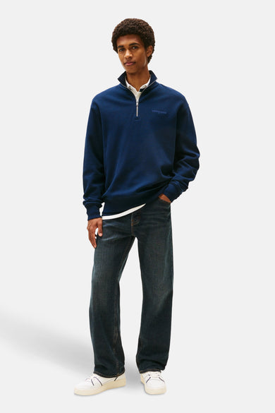 Pull-over bleu de TOMMY JEANS, avec une fermeture à glissière sur la moitié, porté avec un chemisier blanc et des jeans foncés.