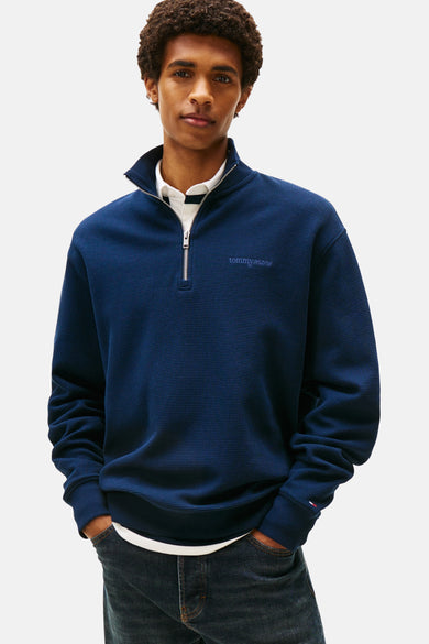 Pull-over bleu de TOMMY JEANS avec fermeture éclair et texture tissée, associé à une chemise blanche et des jeans foncés.