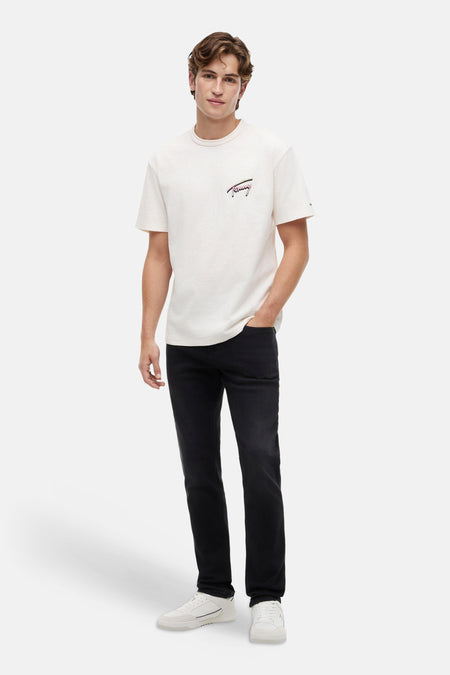 Jean slim noir de TOMMY JEANS, porté par un homme avec un t-shirt de couleur crème et des baskets blanches.