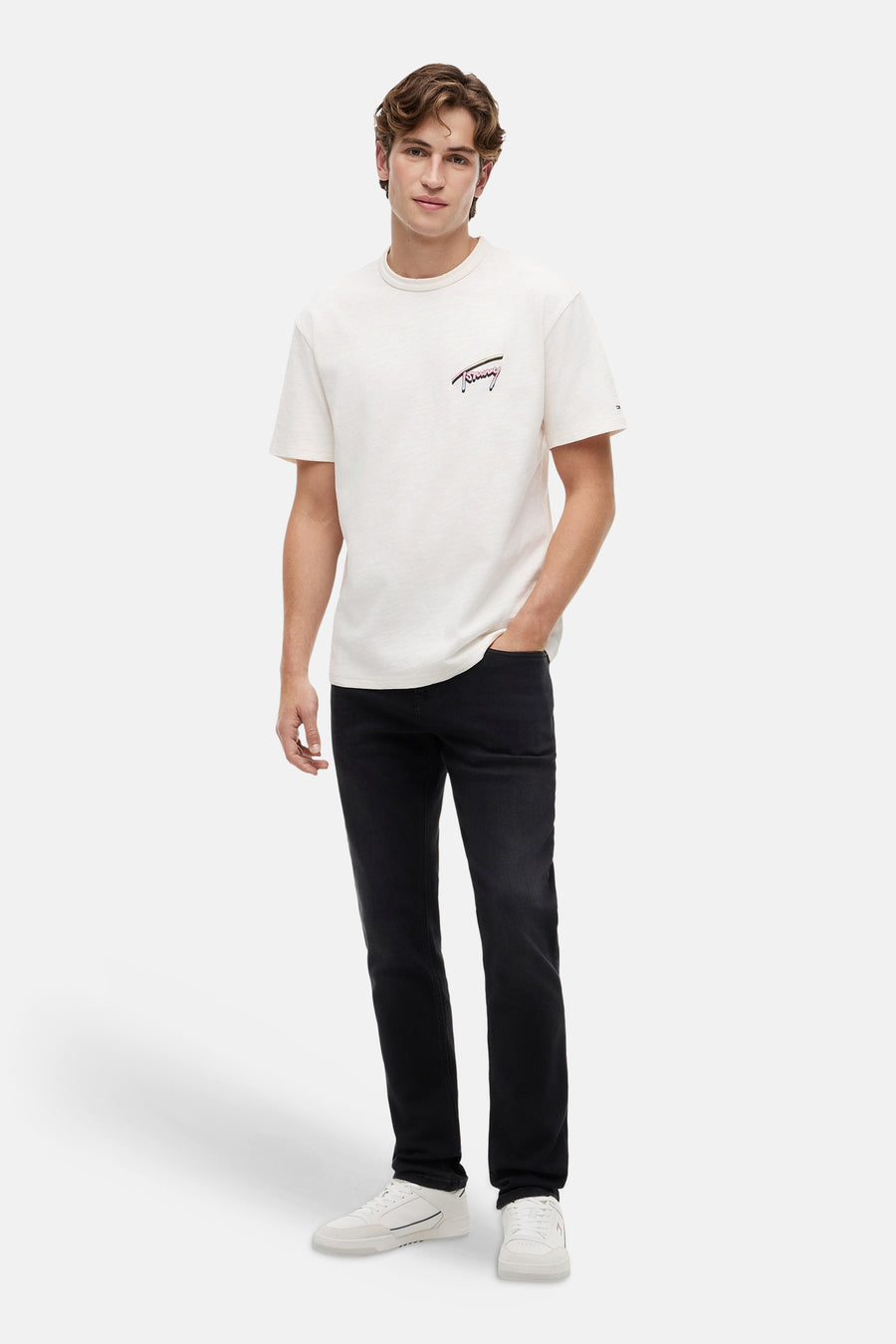 Jean slim noir de TOMMY JEANS, porté par un homme avec un t-shirt de couleur crème et des baskets blanches.