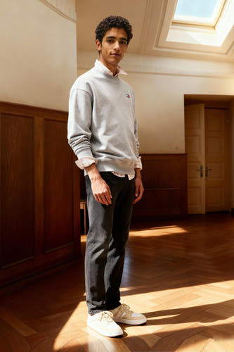 Jean slim noir de TOMMY JEANS, associé à un pull gris et des baskets crème, debout dans une pièce ensoleillée.