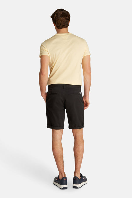 Short noir de TOMMY JEANS, avec des boucles à l'arrière et un bord relevé, porté avec un T-shirt jaune et des baskets bleues.