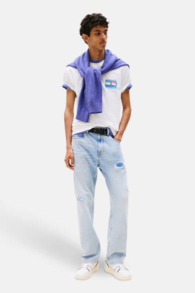 T-shirt blanc à manches courtes de TOMMY JEANS, avec logo sur la poitrine, associé à un pull bleu sur les épaules et des jeans bleu clair.