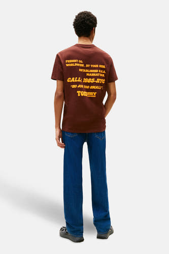 T-shirt brun à manches courtes de TOMMY JEANS, avec une impression textuelle jaune sur le dos, associé à un jean bleu.